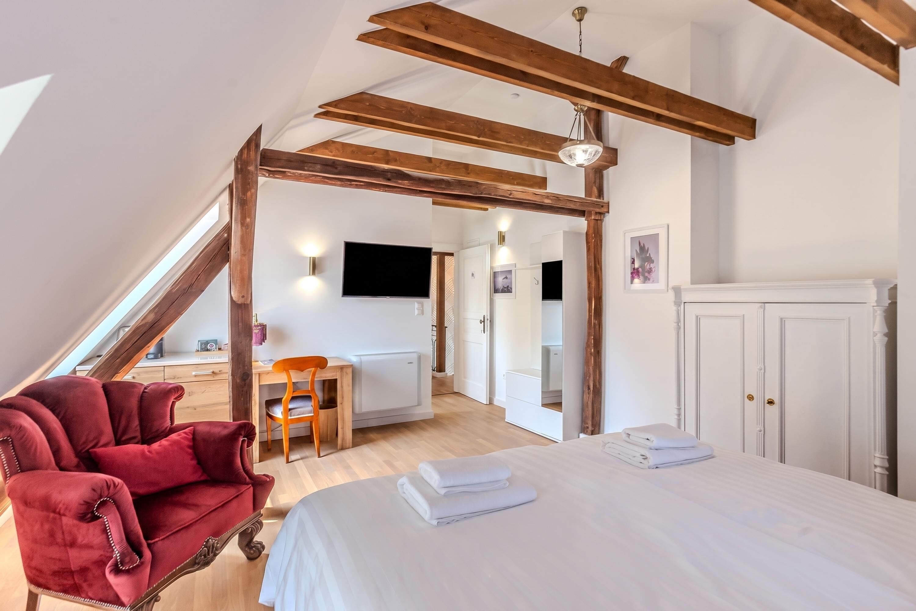 A bedroom with a bed, a red armchair, a wooden ceiling, and a white wardrobe.