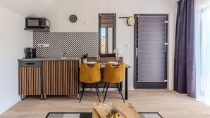 Eine kompakte Küche mit Mikrowelle, Spüle und Holzschränken. Ein Essbereich mit zwei gelben Stühlen und einem Tisch ist ebenfalls zu sehen. Ein Schiebetür und ein Fenster mit Vorhängen sind sichtbar.