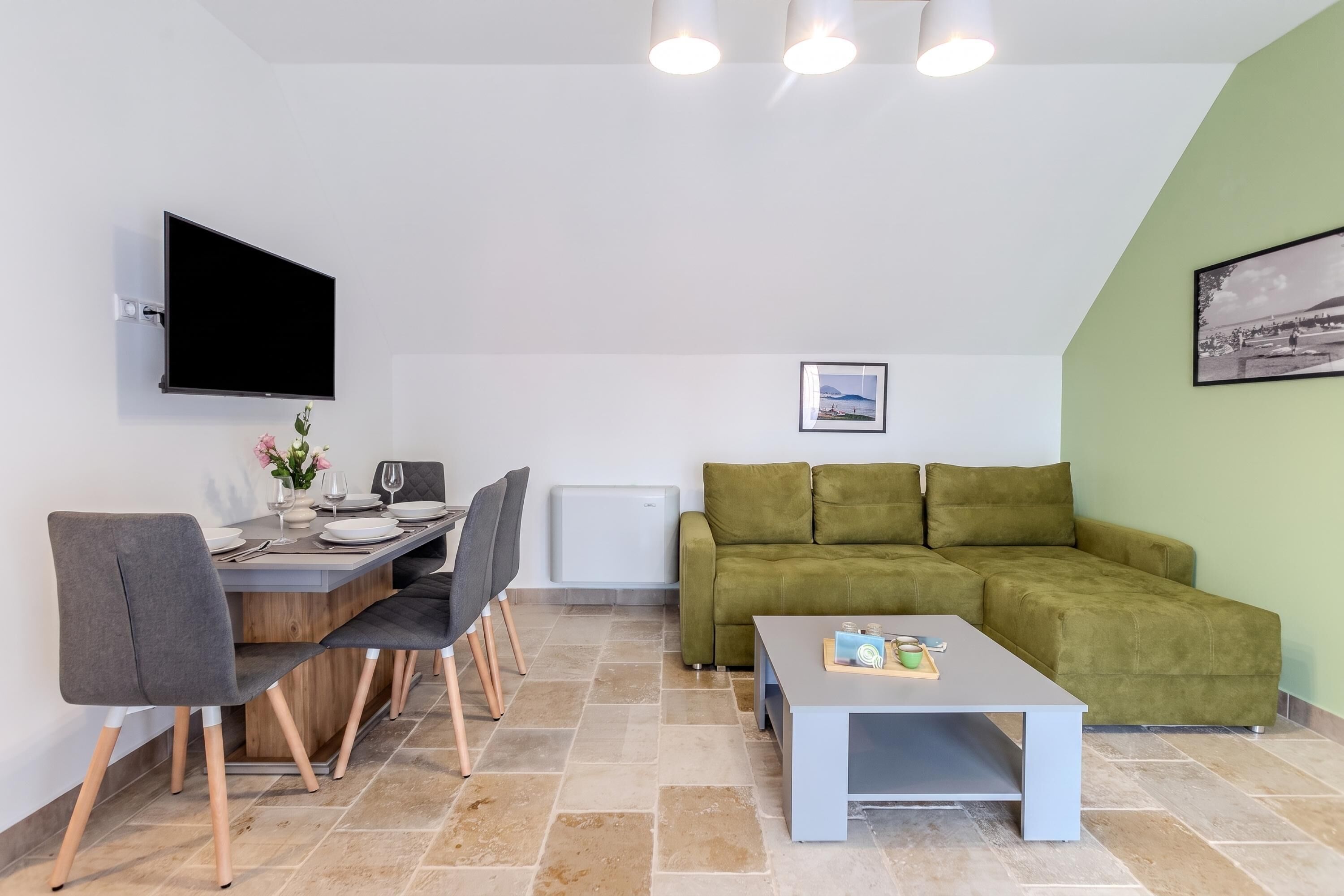 A modern living room with a flat-screen TV, a dining area with a table and chairs, a green sofa, and a tiled floor.