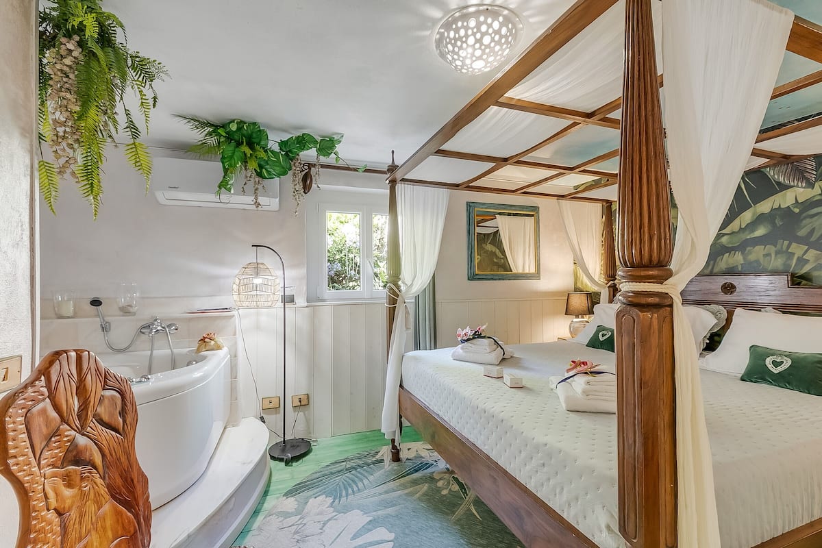 Ein Bett mit Baldachin, ausgestattet mit einer weißen Bettdecke, einem Holz-Kopfende und grünen, herzförmigen Kissen. Im Hintergrund ist eine weiße Badewanne mit Duschvorhang zu sehen.