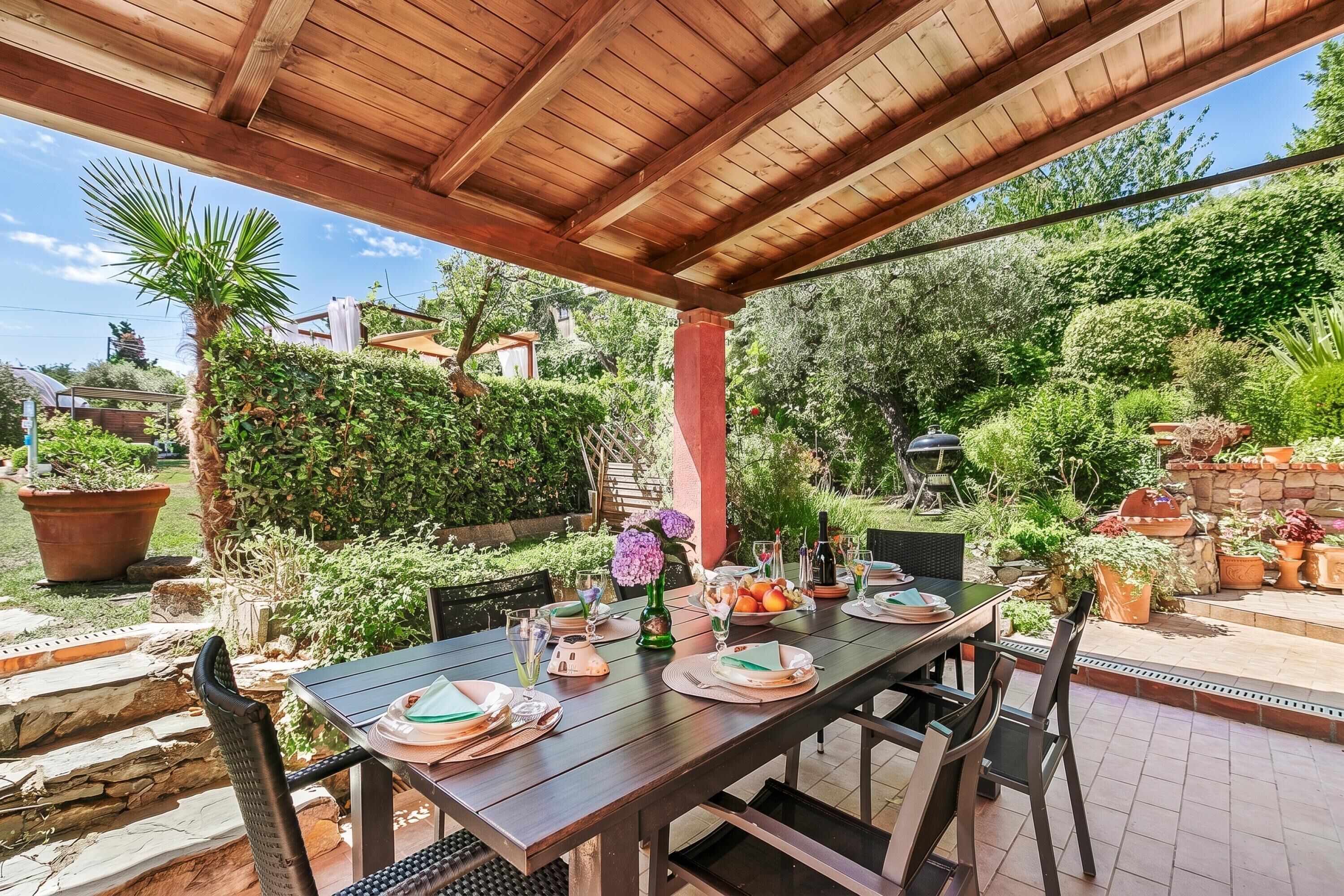 Un espacio exterior techado para comer con una mesa lista para cuatro personas, rodeado de abundante vegetación y plantas en macetas.