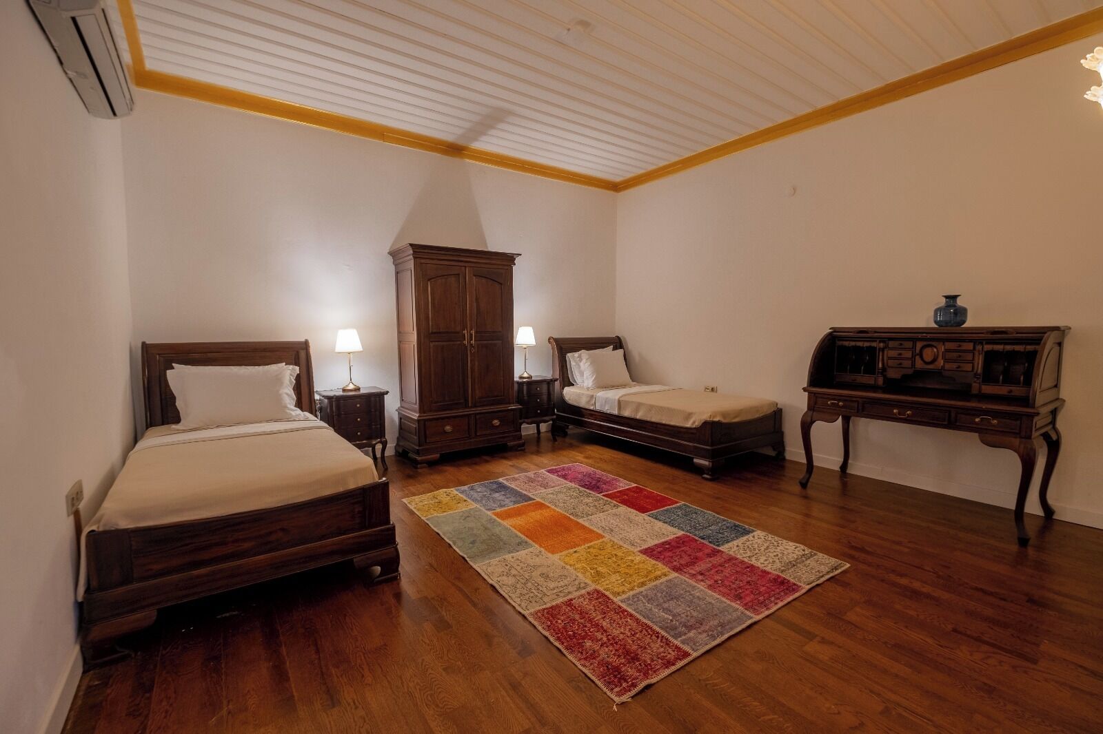 A bedroom with two wooden beds, a large wardrobe, a small table, and a colorful rug.