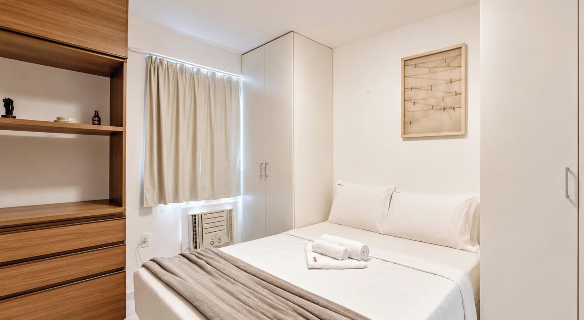 A hotel room with a bed, a window with curtains, a wooden shelf, a radiator, and a wall-mounted picture.