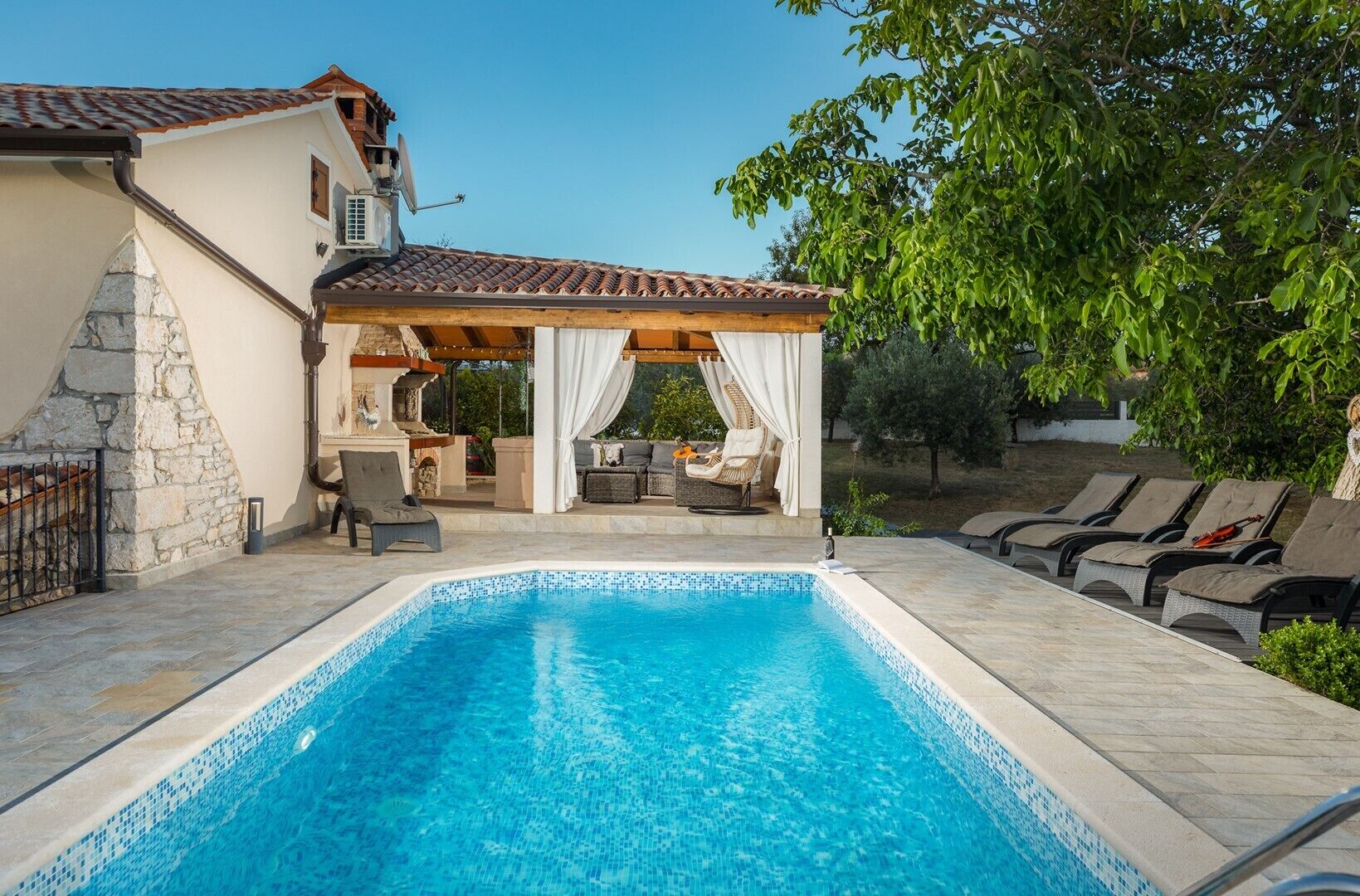A swimming pool with a stone deck, a covered outdoor seating area, and a stone wall.