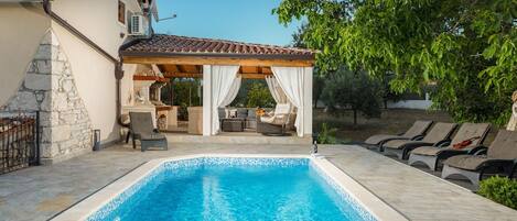 A swimming pool with a stone deck, a covered outdoor seating area, and a stone wall.