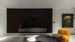 Una sala moderna con una televisión de pantalla plana montada en una pared de madera oscura, una mesa de centro redonda, un sillón amarillo mostaza y un otoman blanco.