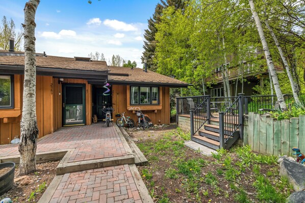 A wooden house with a brick pathway leading to the entrance, a wooden deck, and a fenced garden area.