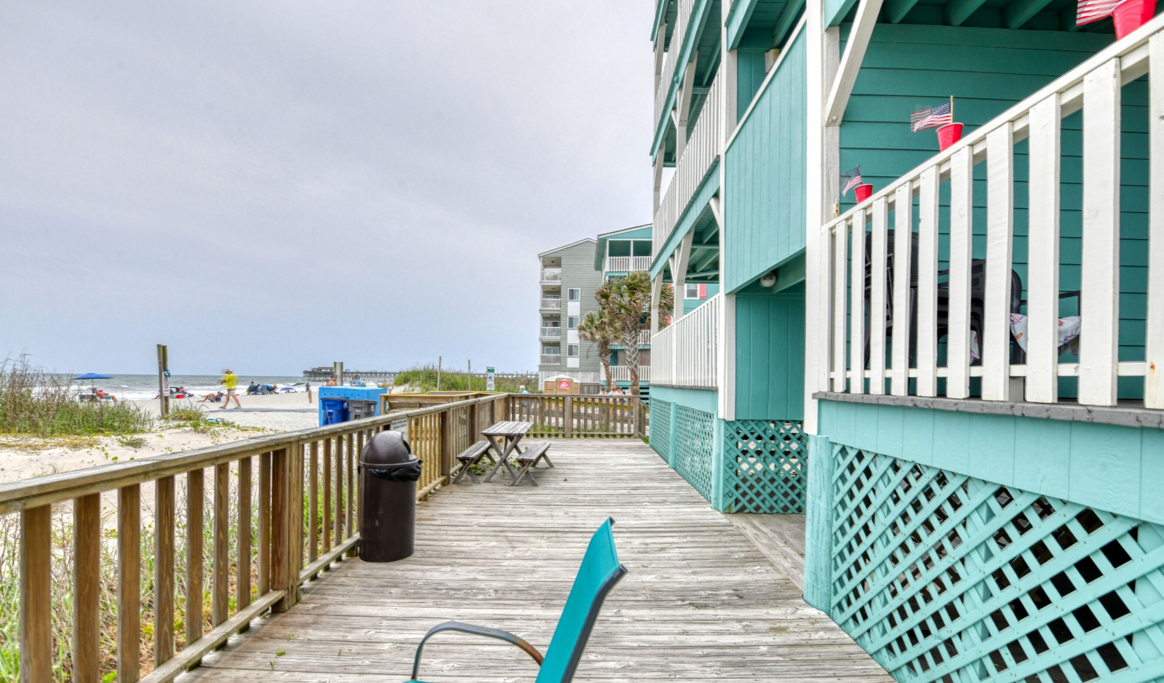 A wooden deck with a view of the beach and ocean, a trash can, and a table with chairs.