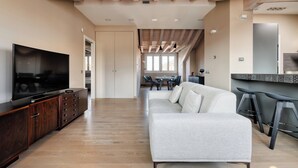 Un salon moderne doté d’un téléviseur à écran plat, d’un meuble de rangement en bois foncé, d’un canapé gris clair et d’un coin cuisine avec un réfrigérateur et des tabourets de bar.
