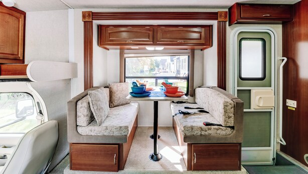 A compact RV interior with a kitchenette, a dining area with two cushioned seating, and a window providing a view of the outside. - Custom Motorhome that brings joy. (Ann Arbor)