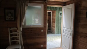 A wooden cabin room with a bed, a chair, a window with curtains, and a door leading to a hallway.