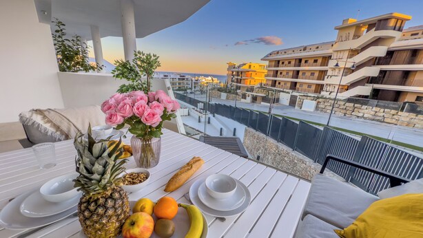 A balcony with a table set for breakfast, featuring fruits, bread, and a vase of flowers.