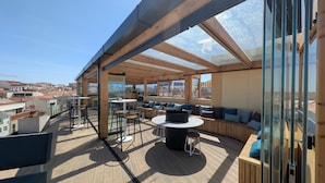 Eine Dachterrasse mit Holzboden, einem runden Tisch, Sitzbereichen und Glaswänden mit Blick auf die Stadt.