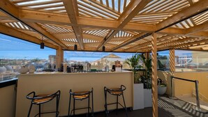 Eine Dachterrasse mit Holzbalken, drei Barhocker, einer Theke und Blick auf die Stadt.