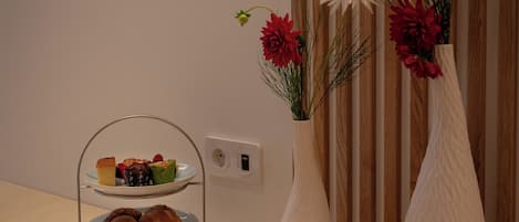 A table with a tiered dessert stand, two white vases with flowers, and a power outlet on a wooden wall.