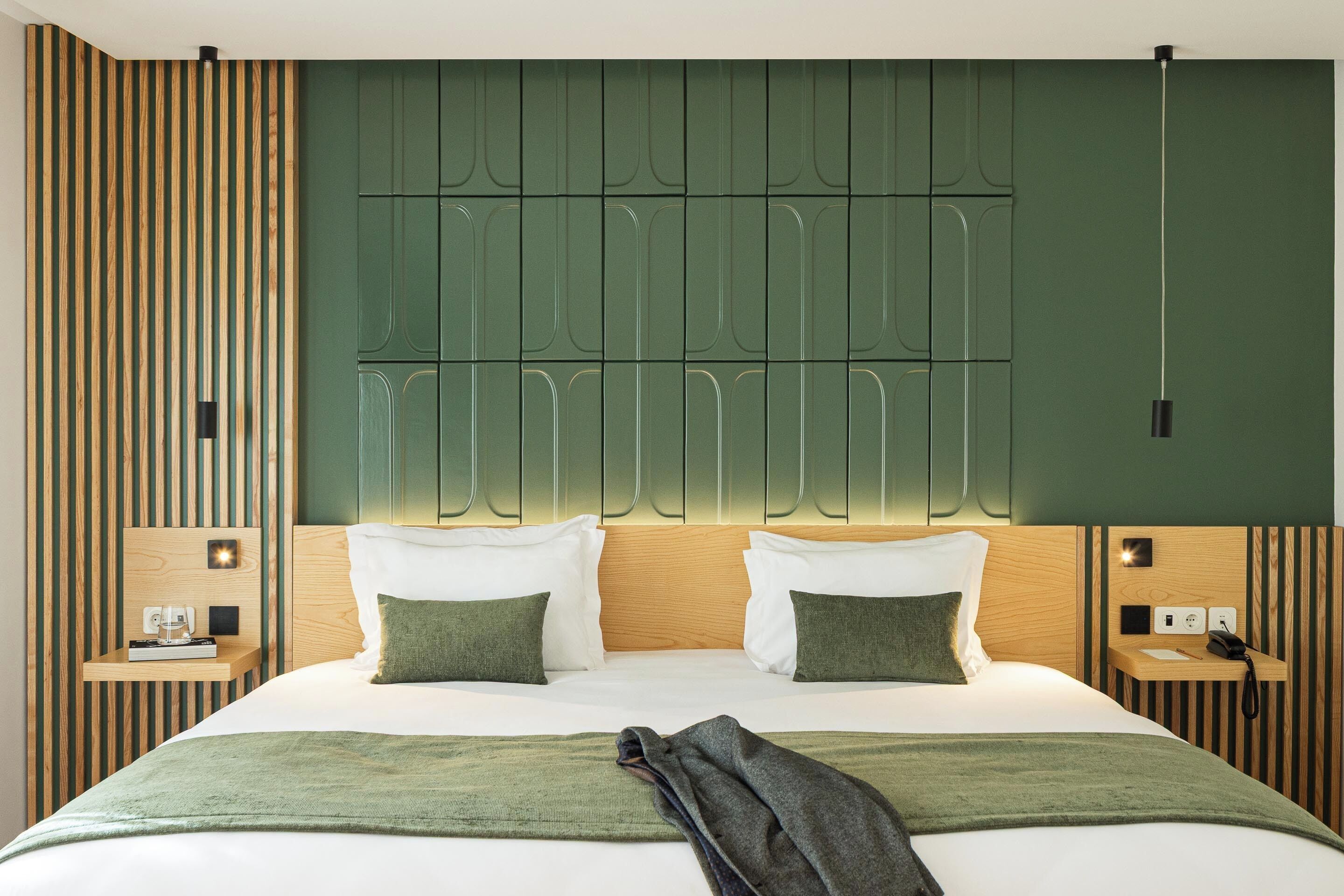 A modern hotel room with a large bed, two bedside tables, a headboard with built-in lighting, and a green accent wall.
