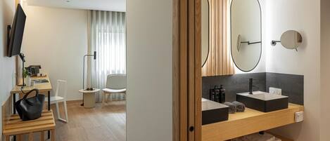 Une chambre d’hôtel moderne, dotée d’un bureau en bois, d’une chaise, d’une télévision, d’un grand miroir et d’une salle de bain avec un lavabo et un porte-serviettes.