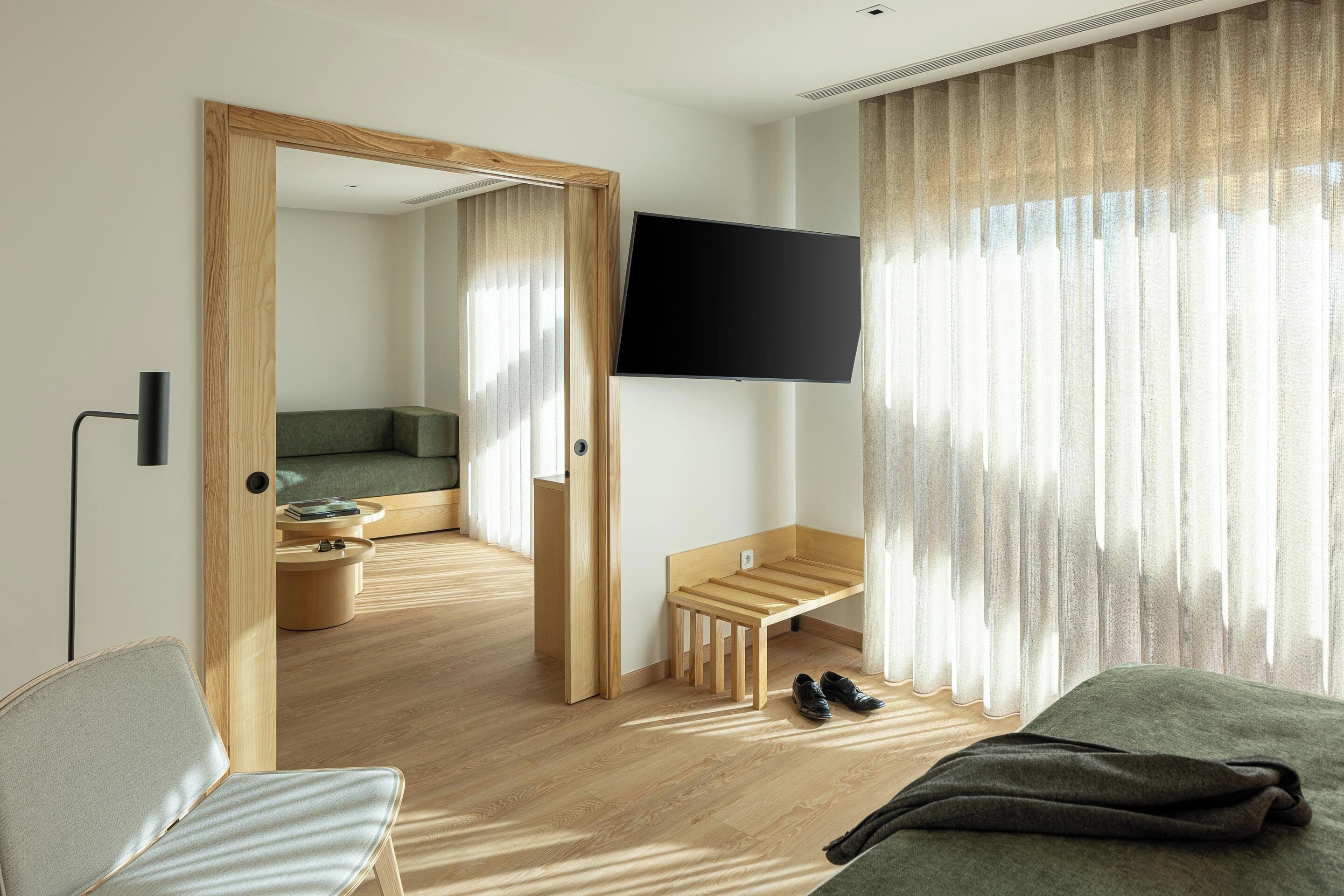 A modern hotel room with a wooden floor, a green sofa, a wooden bench, a TV mounted on the wall, and a large window with sheer curtains.
