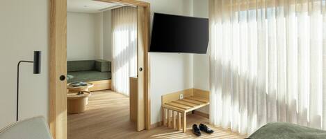 A modern hotel room with a wooden floor, a green sofa, a wooden bench, a TV mounted on the wall, and a large window with sheer curtains.