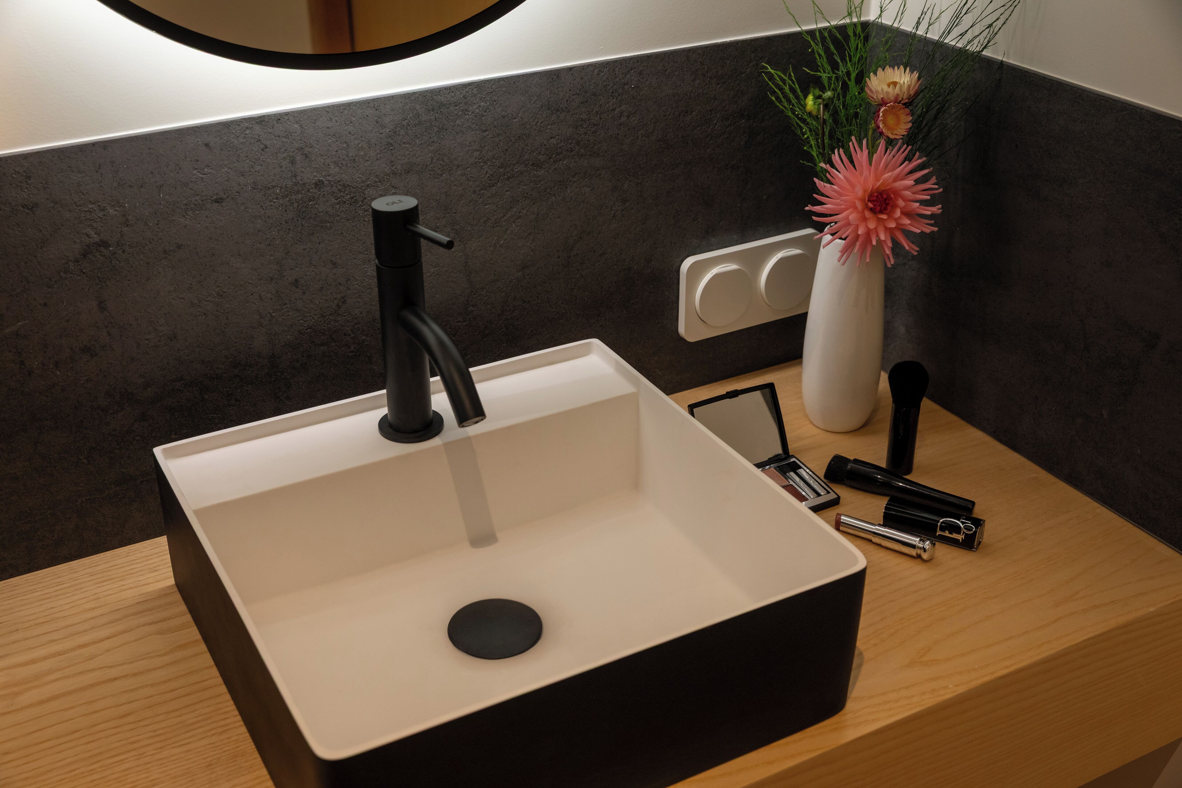 A modern bathroom sink with a black faucet, a white vase with flowers, and a black razor on a wooden countertop.