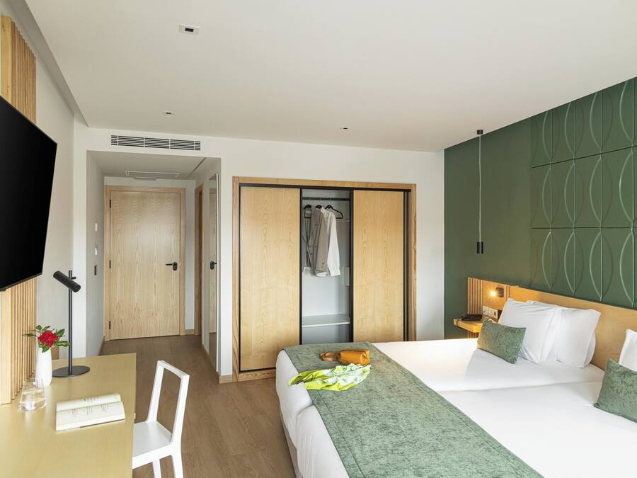Quarto de hotel com uma cama, uma escrivaninha com cadeira, uma televisão e um guarda-roupa.