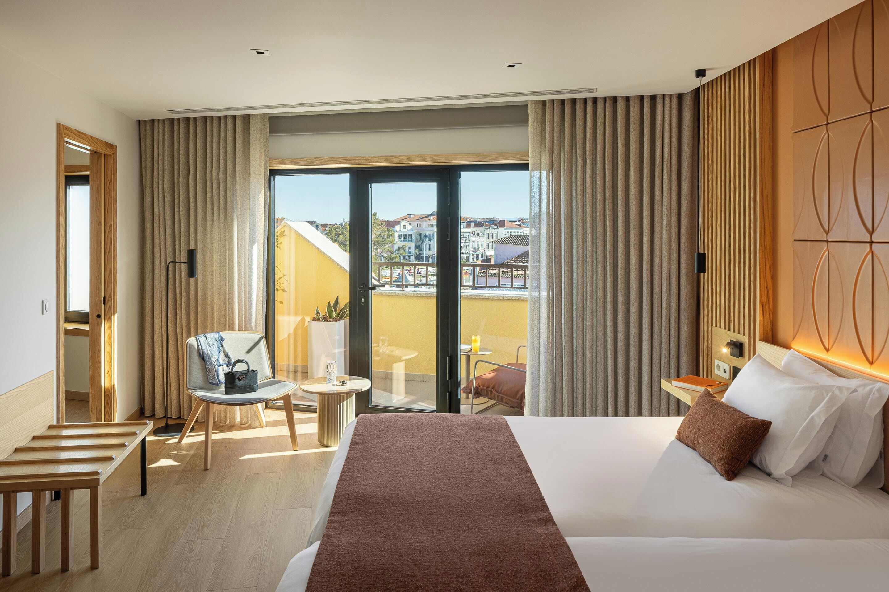 A hotel room with a large bed, a chair, a small table, and a balcony with a view of buildings.