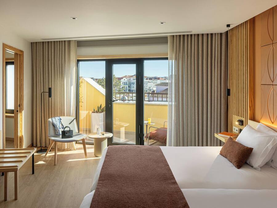 Ein Hotelzimmer mit einem großen Bett, einem Sessel, einem kleinen Tisch und einem Balkon mit Blick auf Gebäude.