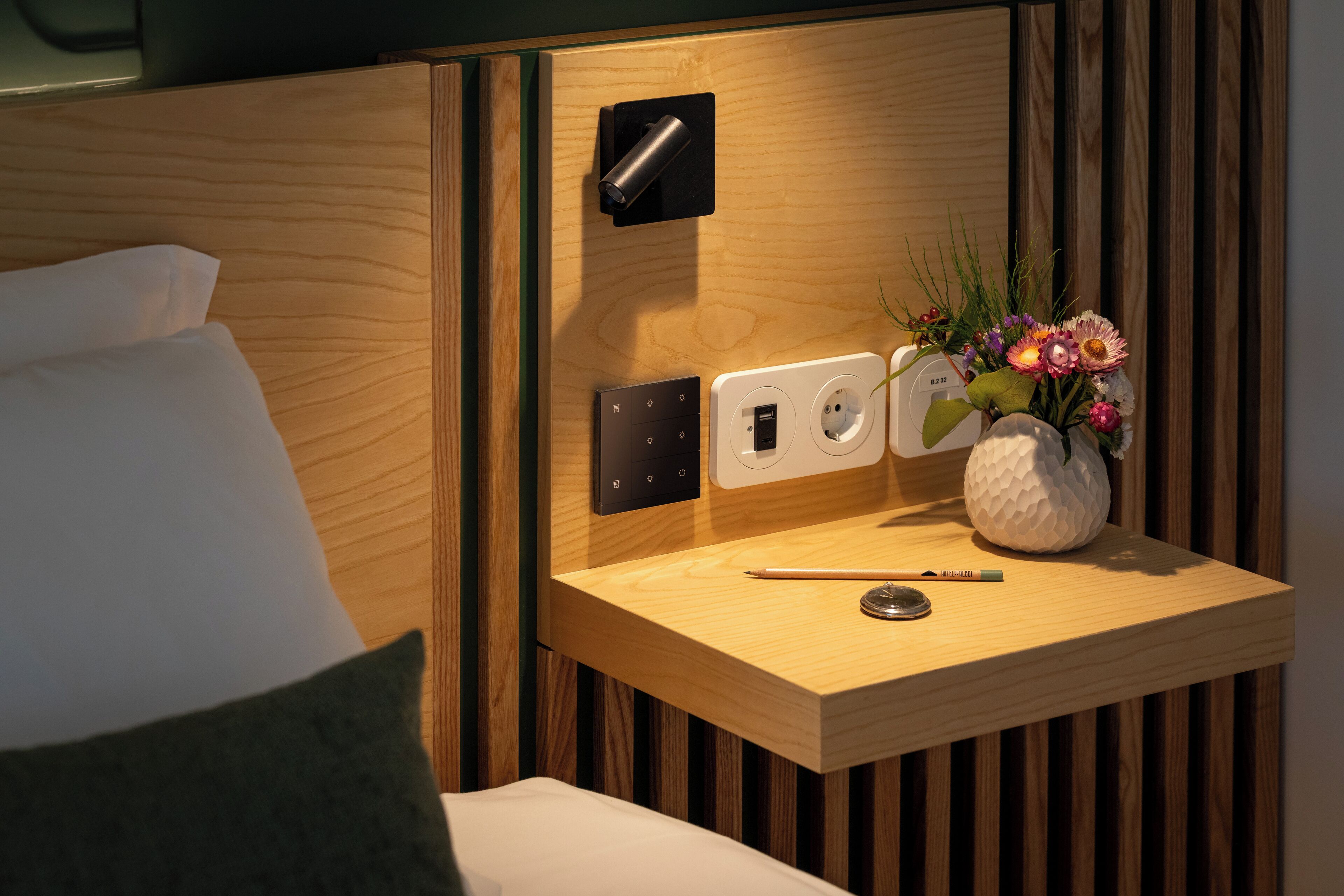 A neatly arranged hotel room with a wooden bedside table, a power outlet, a lamp, and a vase with flowers.