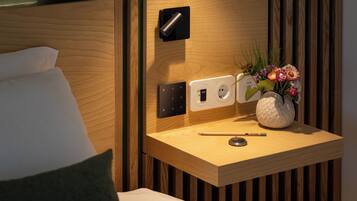 A neatly arranged hotel room with a wooden bedside table, a power outlet, a lamp, and a vase with flowers.
