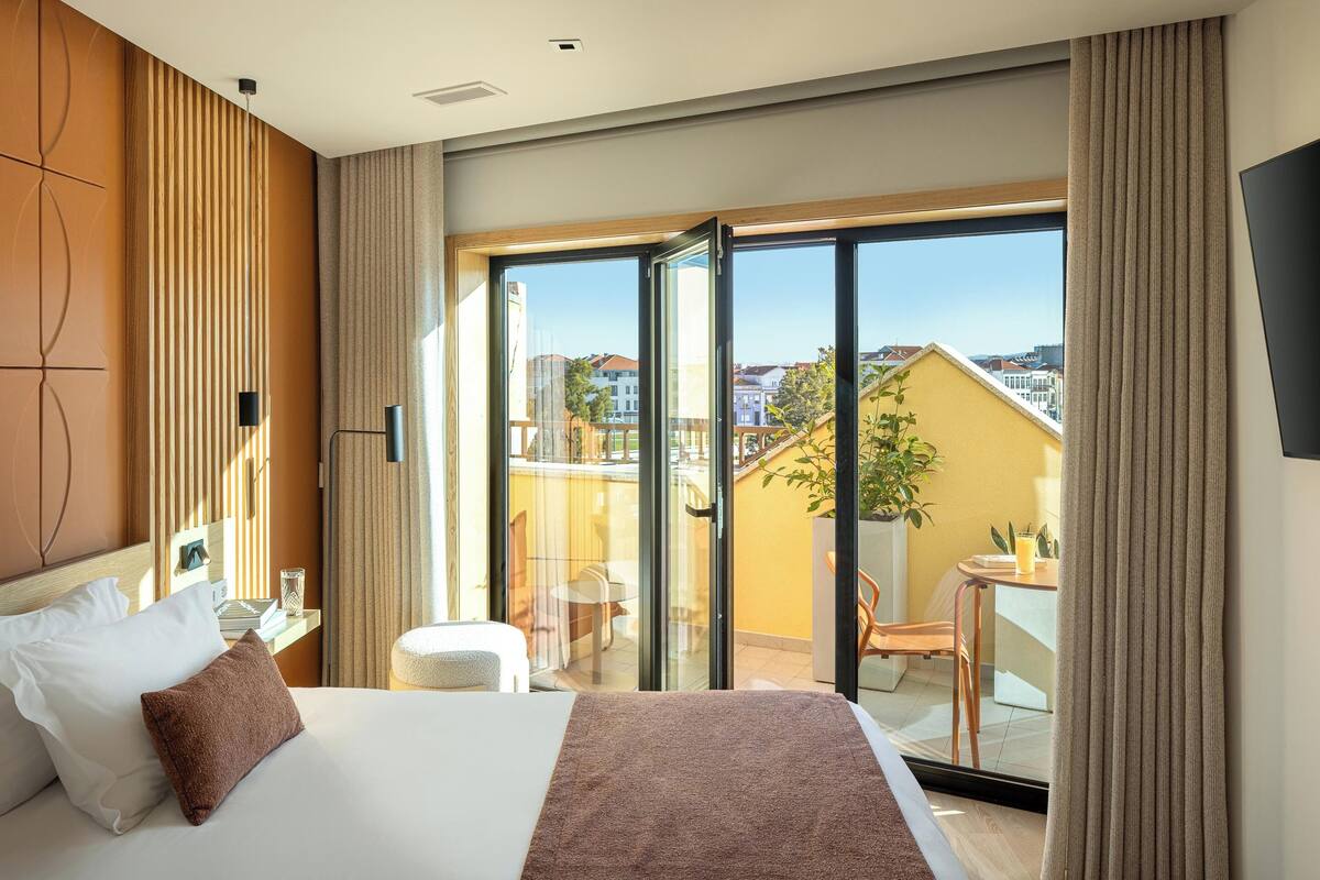 A hotel room with a bed, a desk, a chair, a small table, a potted plant, and a balcony with a view of buildings.