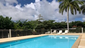 A rectangular swimming pool with clear blue water, surrounded by a metal fence. There are lounge chairs along the poolside, and a palm tree is visible in the background. - Charming studio in the very pleasant district of Orient Bay (SAINT MARTIN)