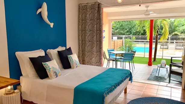 A hotel room with a bed, a blue accent wall, a decorative fish sculpture, a view of a pool and outdoor seating area. - Charming studio in the very pleasant district of Orient Bay (SAINT MARTIN)