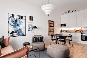 Una sala moderna con sofá, reposapiés y mesa de centro, un comedor con sillas y una mesa, una cocina con gabinetes y electrodomésticos blancos, y obras de arte en las paredes.
