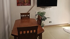 A dining area with a wooden table and chairs, a flat-screen TV mounted on the wall, a painting above the TV, and a potted plant beside the TV.