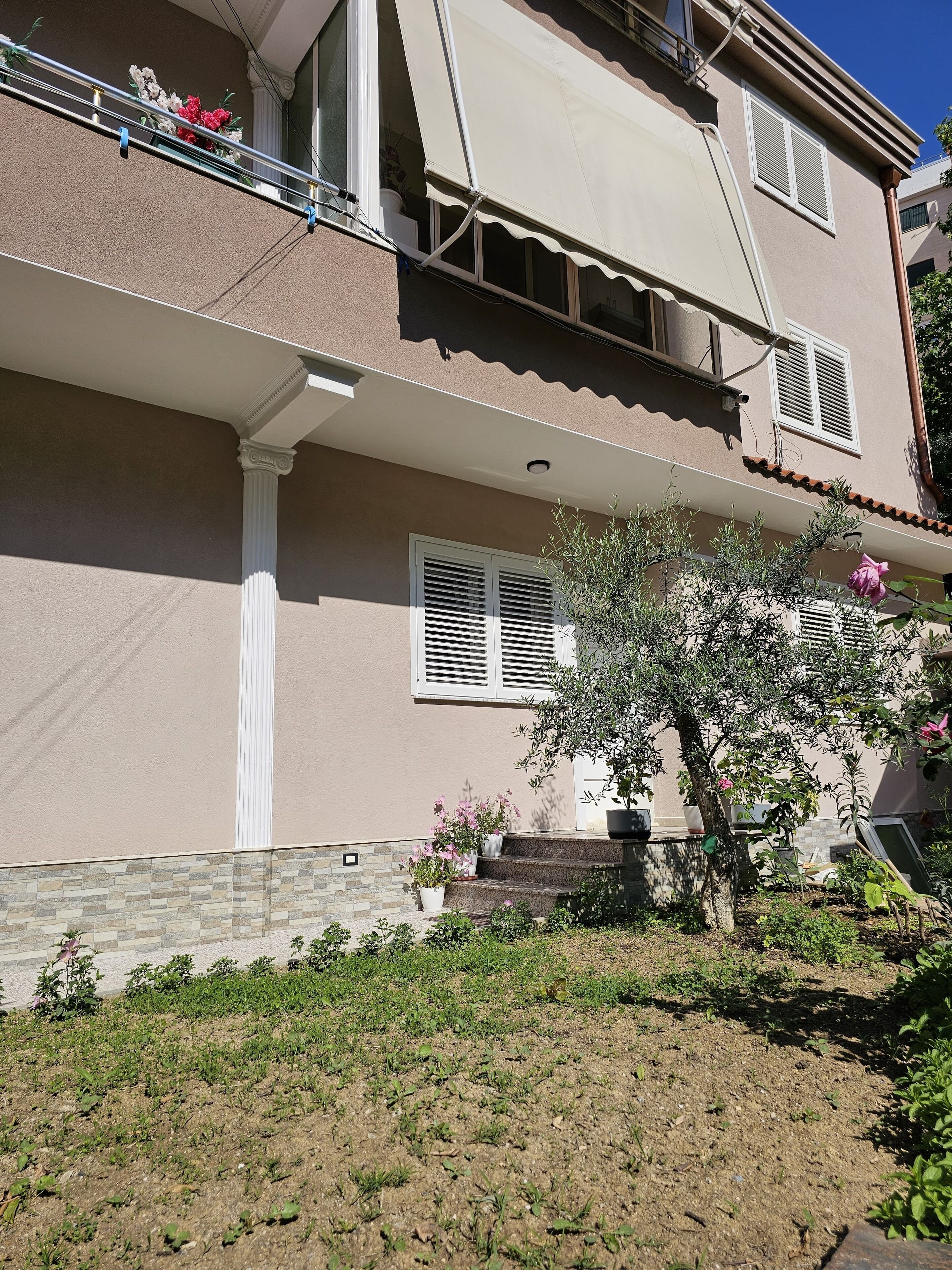 Ein zweistöckiges Gebäude mit Balkon, überdachter Veranda und einem Gartenbereich mit Blumentöpfen und einem kleinen Baum.