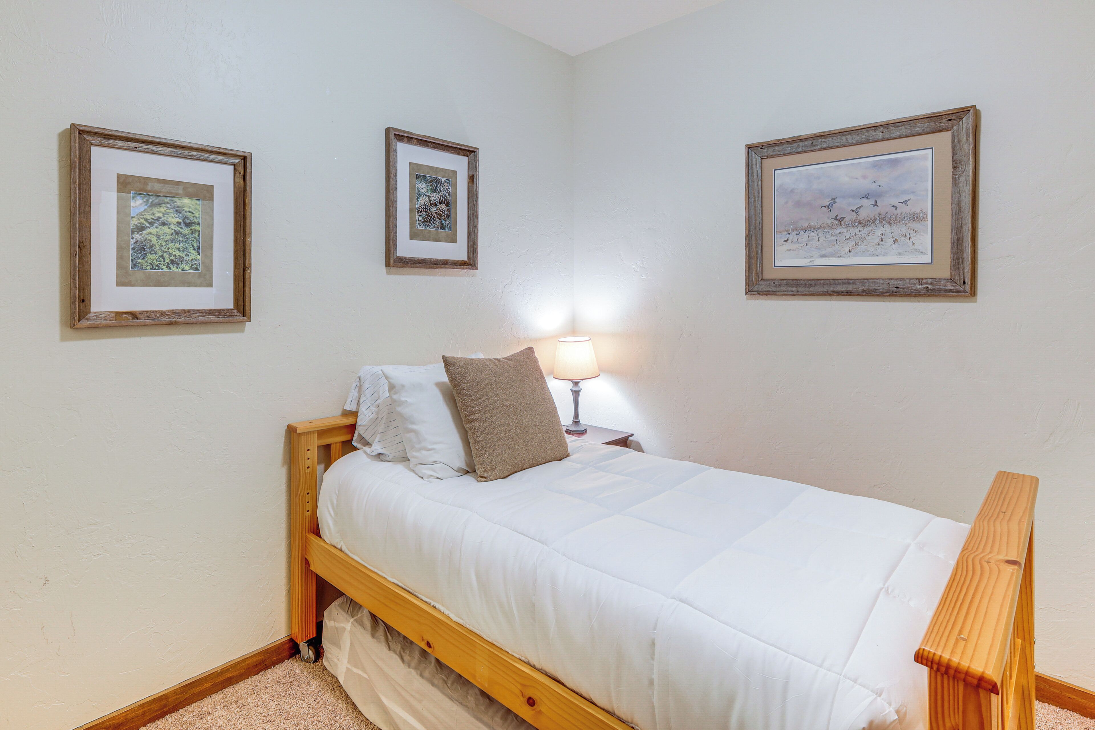 A bedroom with a wooden bed, white bedding, two framed pictures on the wall, and a bedside lamp.