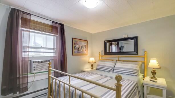 Quarto de hotel com uma cama, mesas de cabeceira, janela com cortinas, unidade de ar condicionado, espelho fixado na parede e uma moldura com uma imagem.