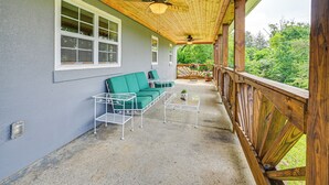 Un porche cubierto con un sofá verde, mesas de metal blanco y una barandilla de madera con vistas a un jardín exuberante.