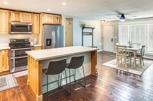 A modern kitchen with wooden cabinets, a stainless steel refrigerator, a microwave, and an oven. There is a bar area with two stools and a dining table with chairs.
