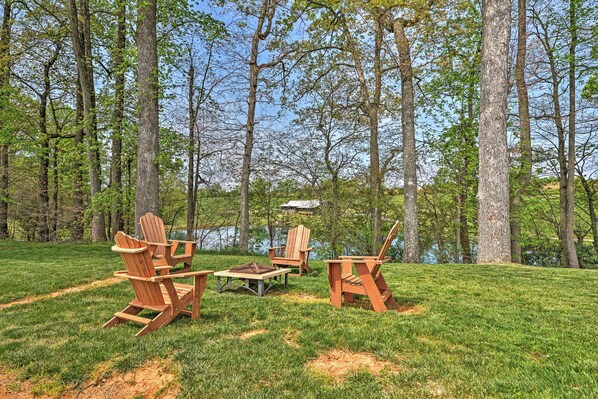 A serene outdoor seating area with Adirondack chairs and a picnic table, surrounded by lush greenery and tall trees. - Dobson Farmhouse w/ Wraparound Porch & Fire Pit! (Elkin)