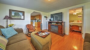 Un salon comprenant un canapé, un pouf et une table basse avec des magazines. On y trouve également un fauteuil à bascule, une cheminée et un téléviseur. Le sol est en bois et le salon est éclairé par un luminaire au plafond.