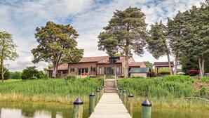 Une jetée en bois mène à une maison dont le toit est en pente, entourée de grands arbres et d’une zone herbeuse.