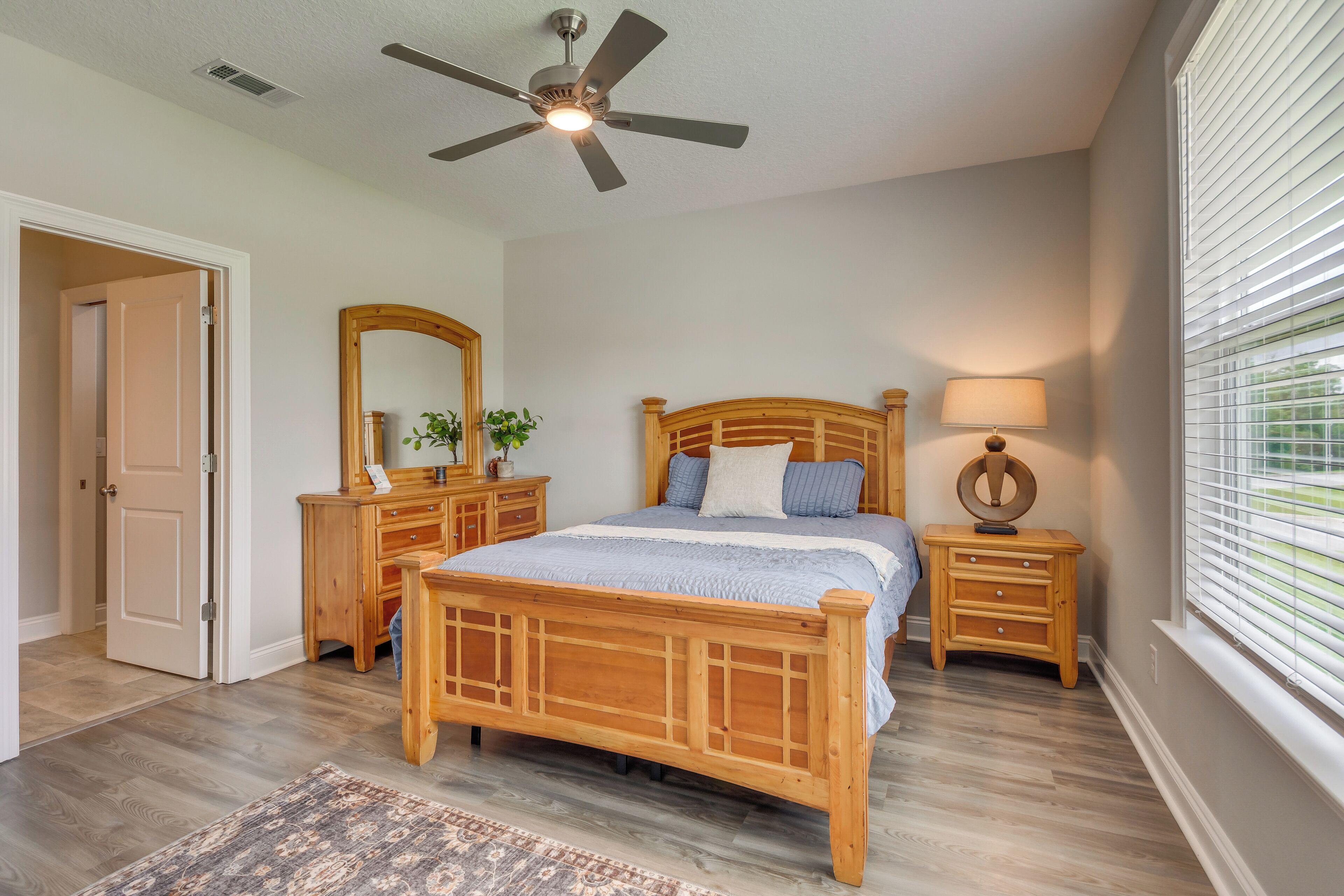 Une chambre à coucher avec un lit en bois, un ventilateur de plafond, une table de chevet avec une lampe, un miroir et une fenêtre avec des stores.