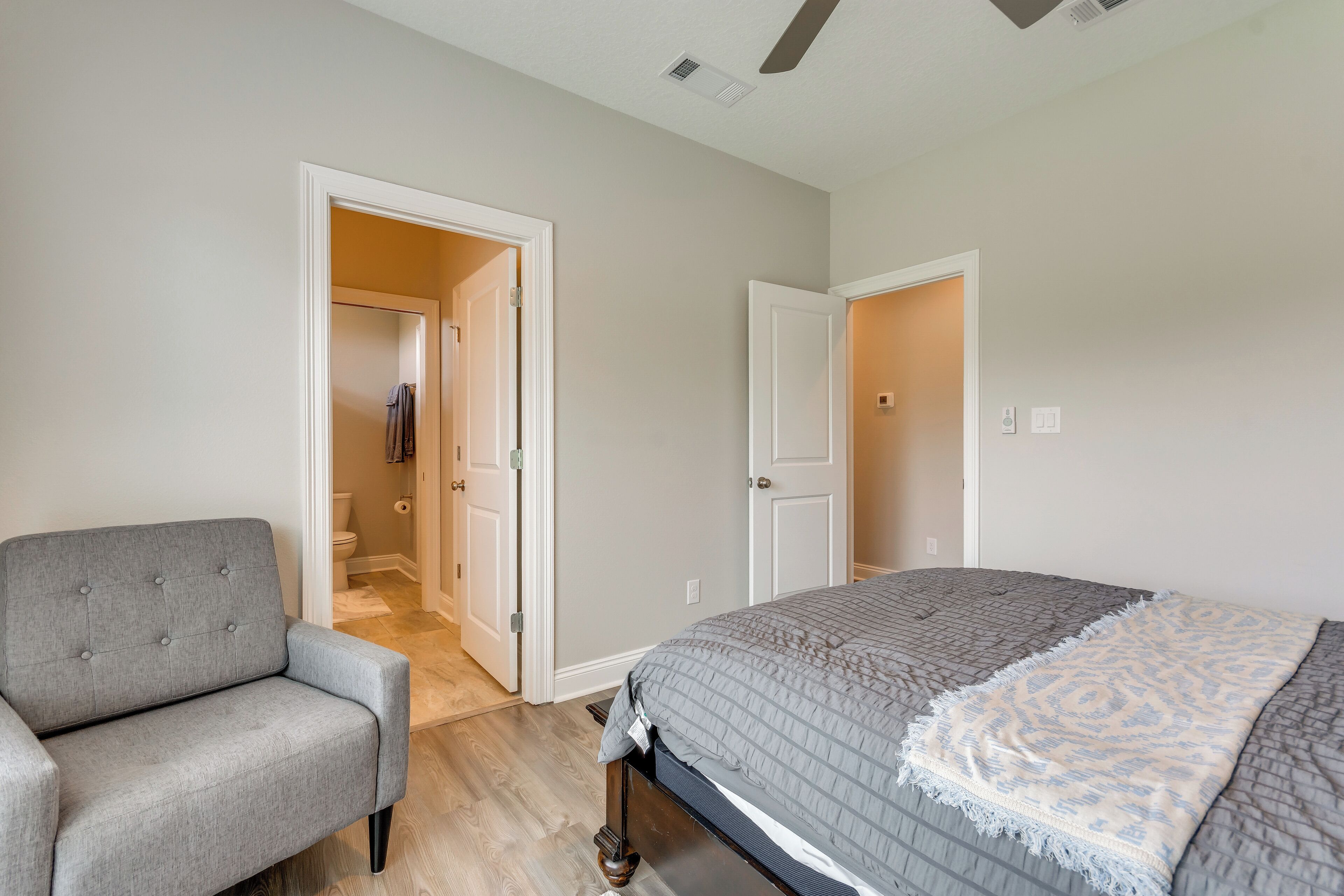 Une chambre à coucher avec un lit, un fauteuil gris et une porte donnant sur une salle de bain.