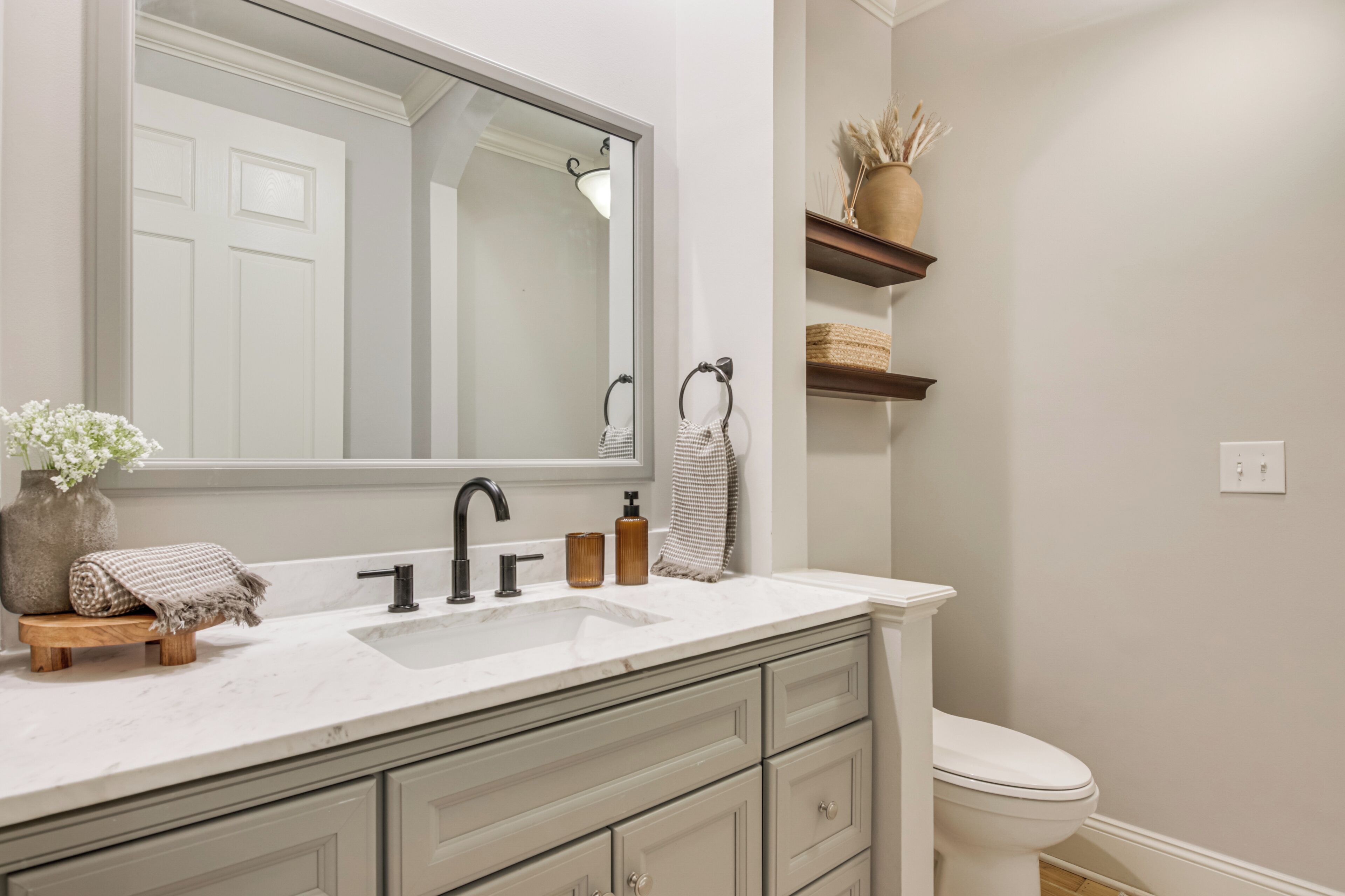 Une salle de bain avec un lavabo blanc, un grand miroir, des toilettes et une étagère ornée d’objets décoratifs.