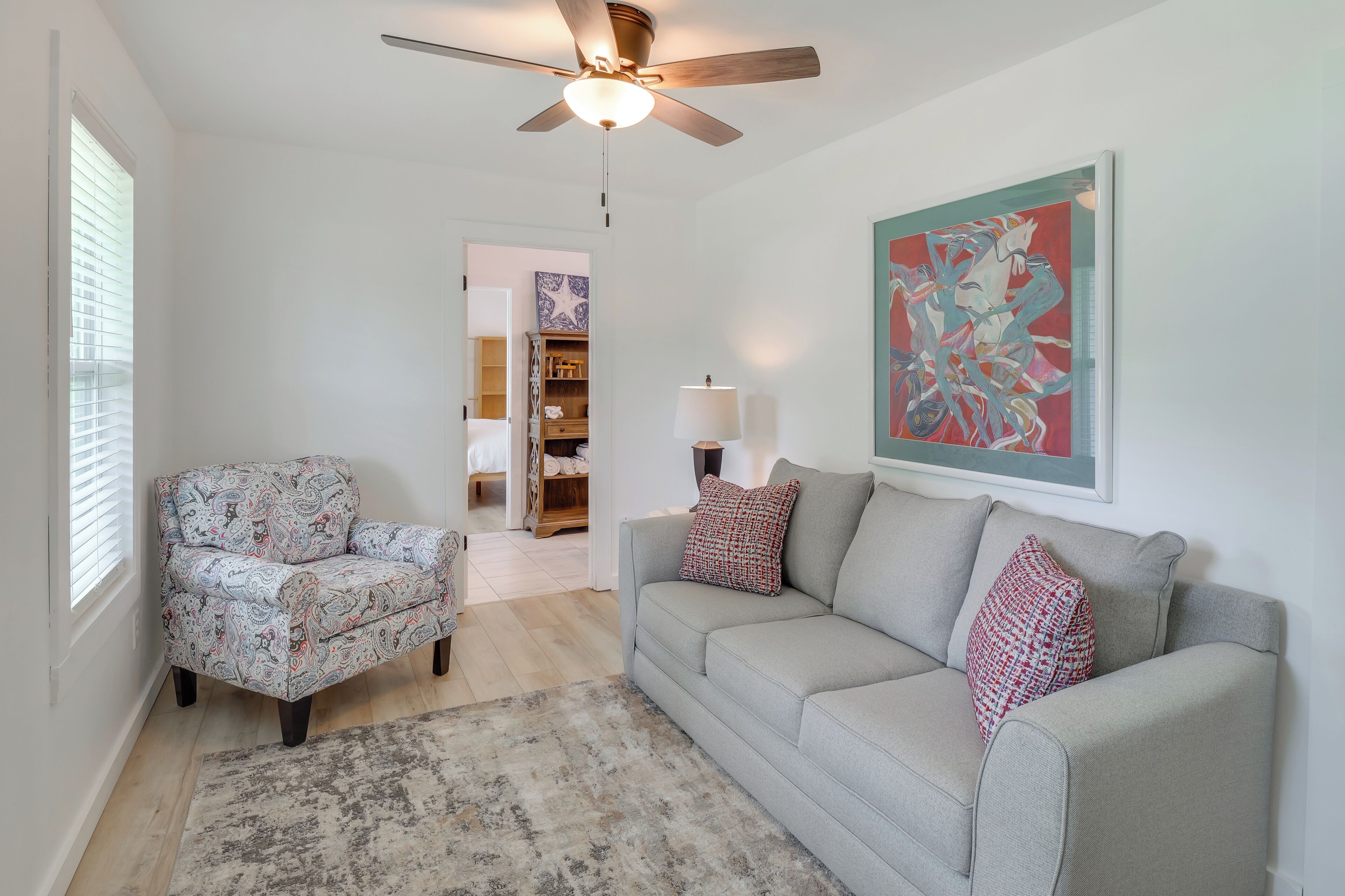 A living room with a ceiling fan, a patterned armchair, a grey sofa with patterned cushions, a rug, and a painting on the wall.