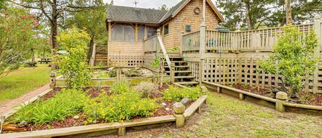 A wooden cabin with a deck, surrounded by a garden with various plants and a wooden fence.