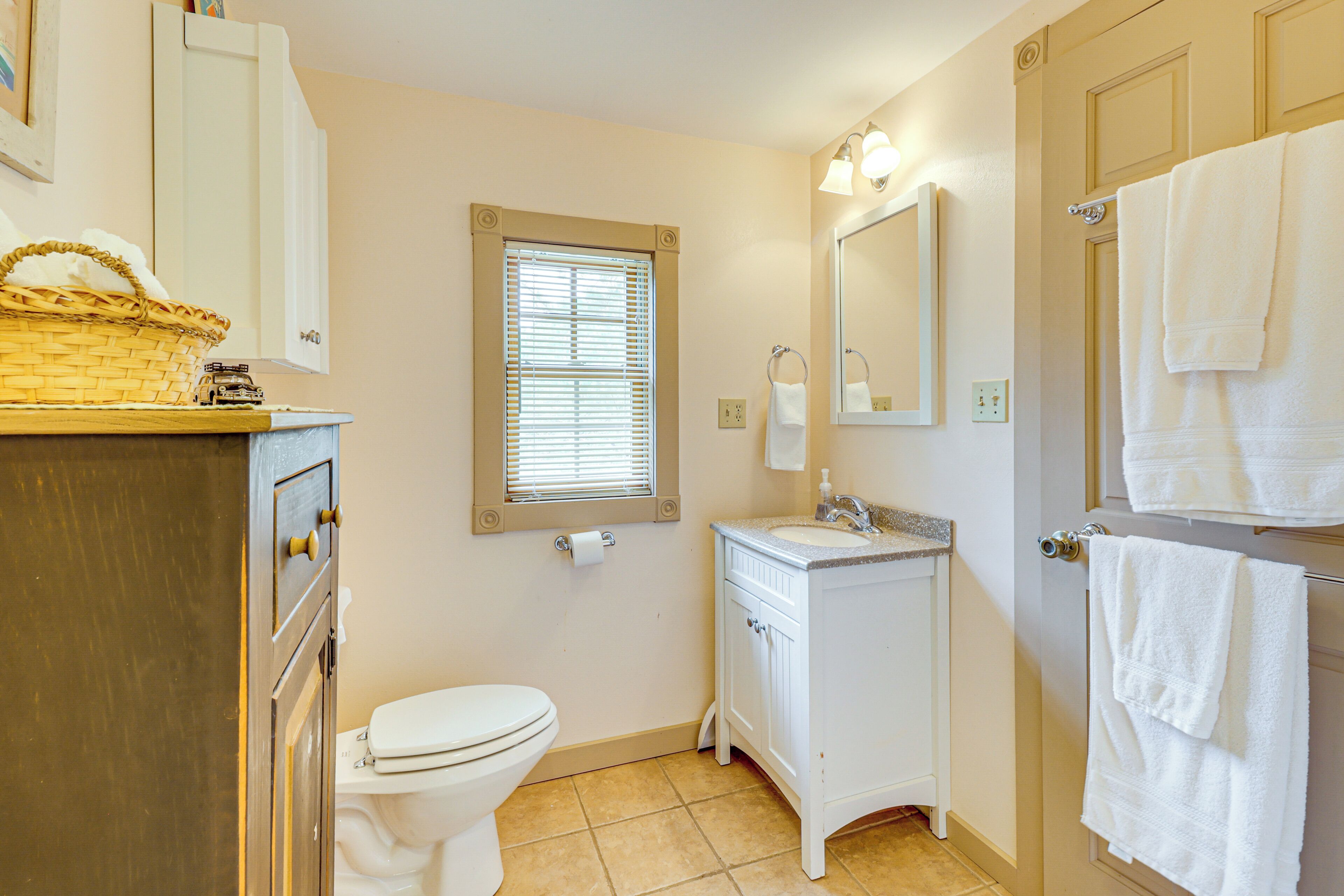 A bathroom with a toilet, sink, and mirror. There is a window with blinds, a towel rack, and a basket on the counter.
