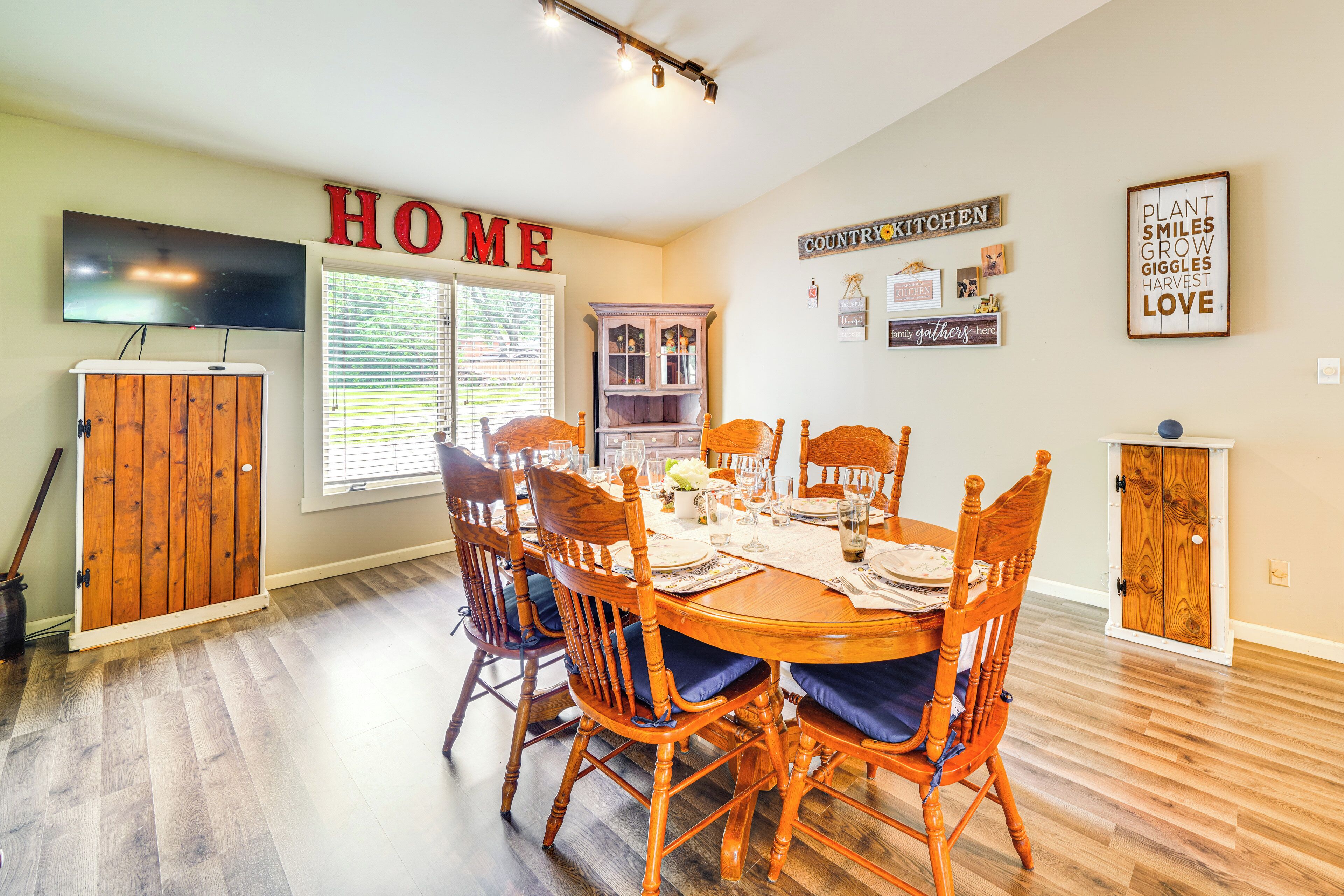 A dining area with a wooden table set for six, surrounded by matching chairs. A television is mounted on the wall above the table.