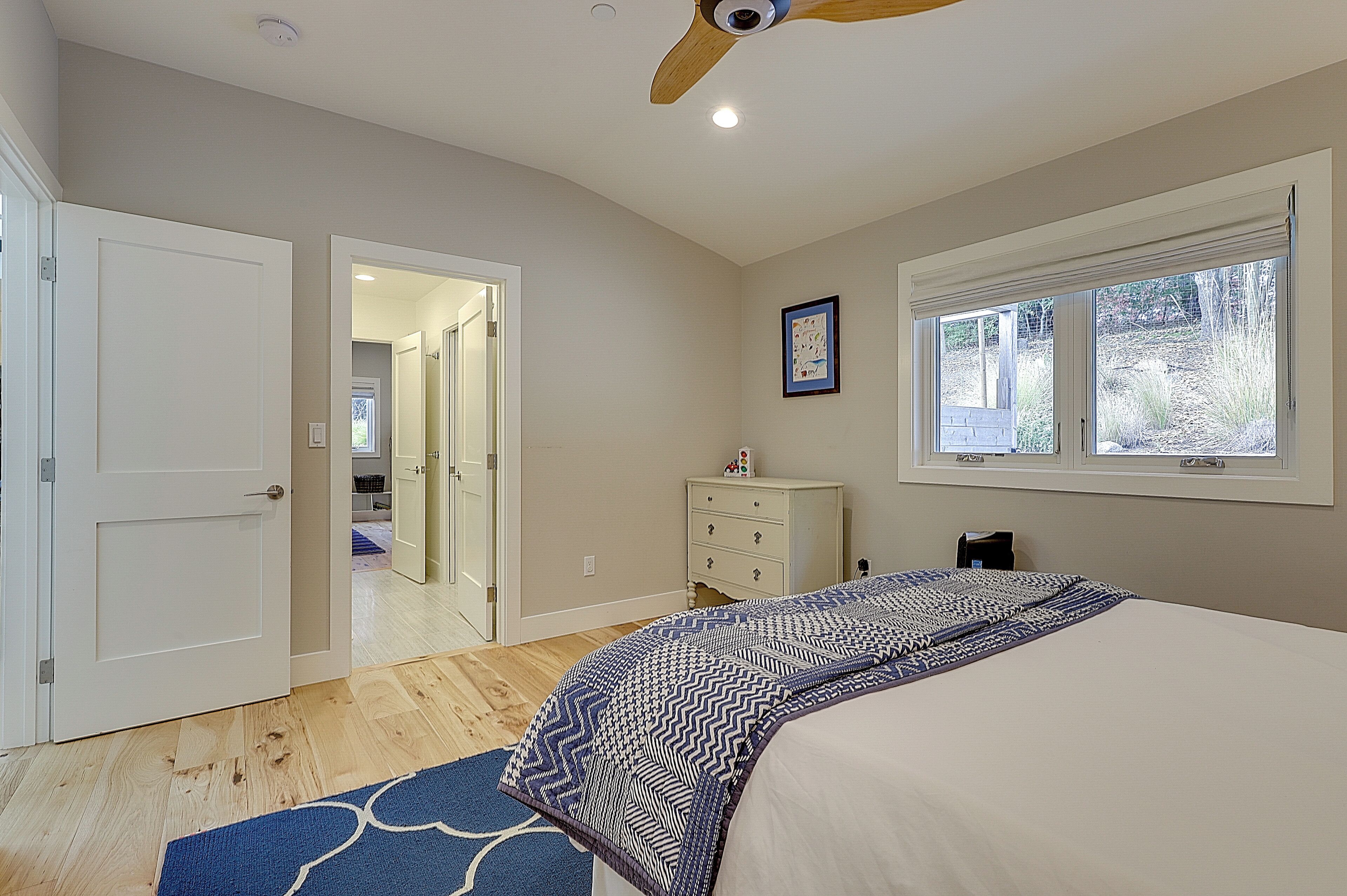 A bedroom with a bed, a dresser, a ceiling fan, a window, and a door leading to another room.